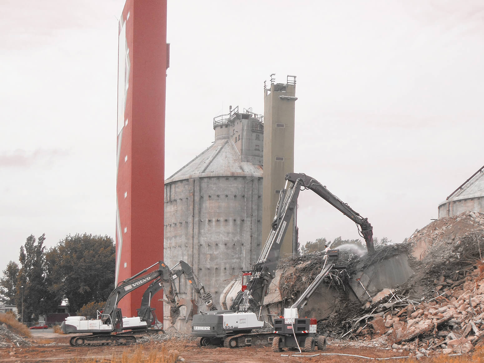 Several heavy construction vehicles tear down buildings made of reinforced concrete with two old digester towers in the background